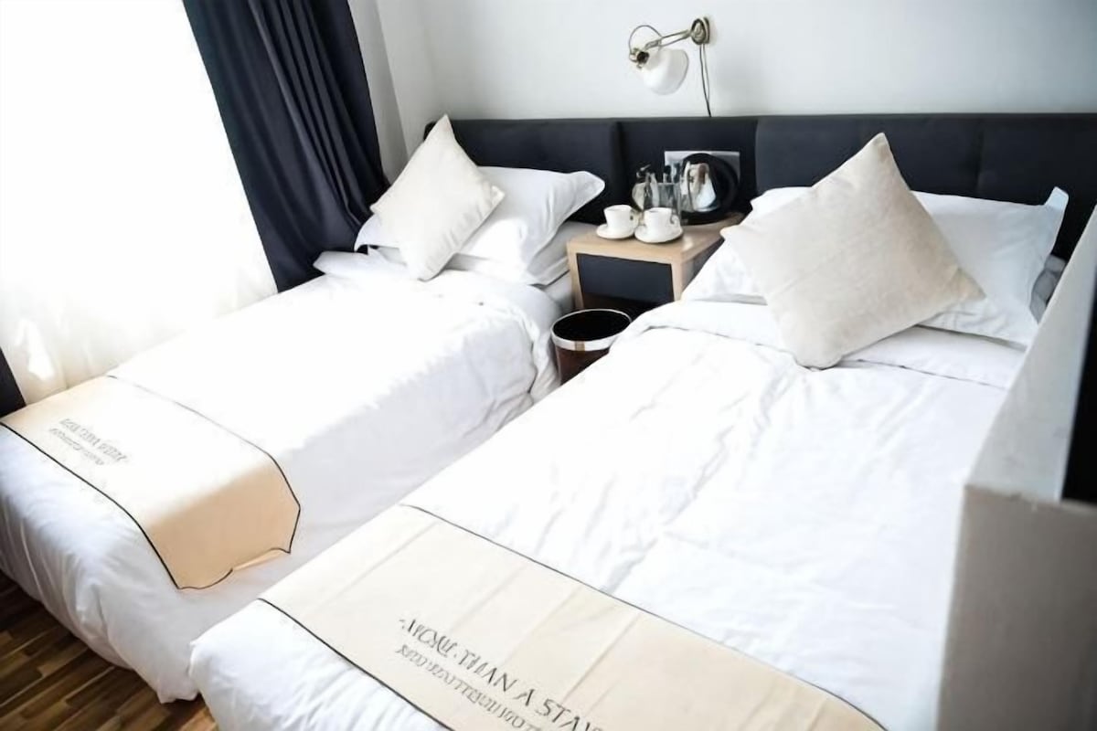 A hotel room with two beds, a nightstand with a lamp, and a view of the outdoors.
