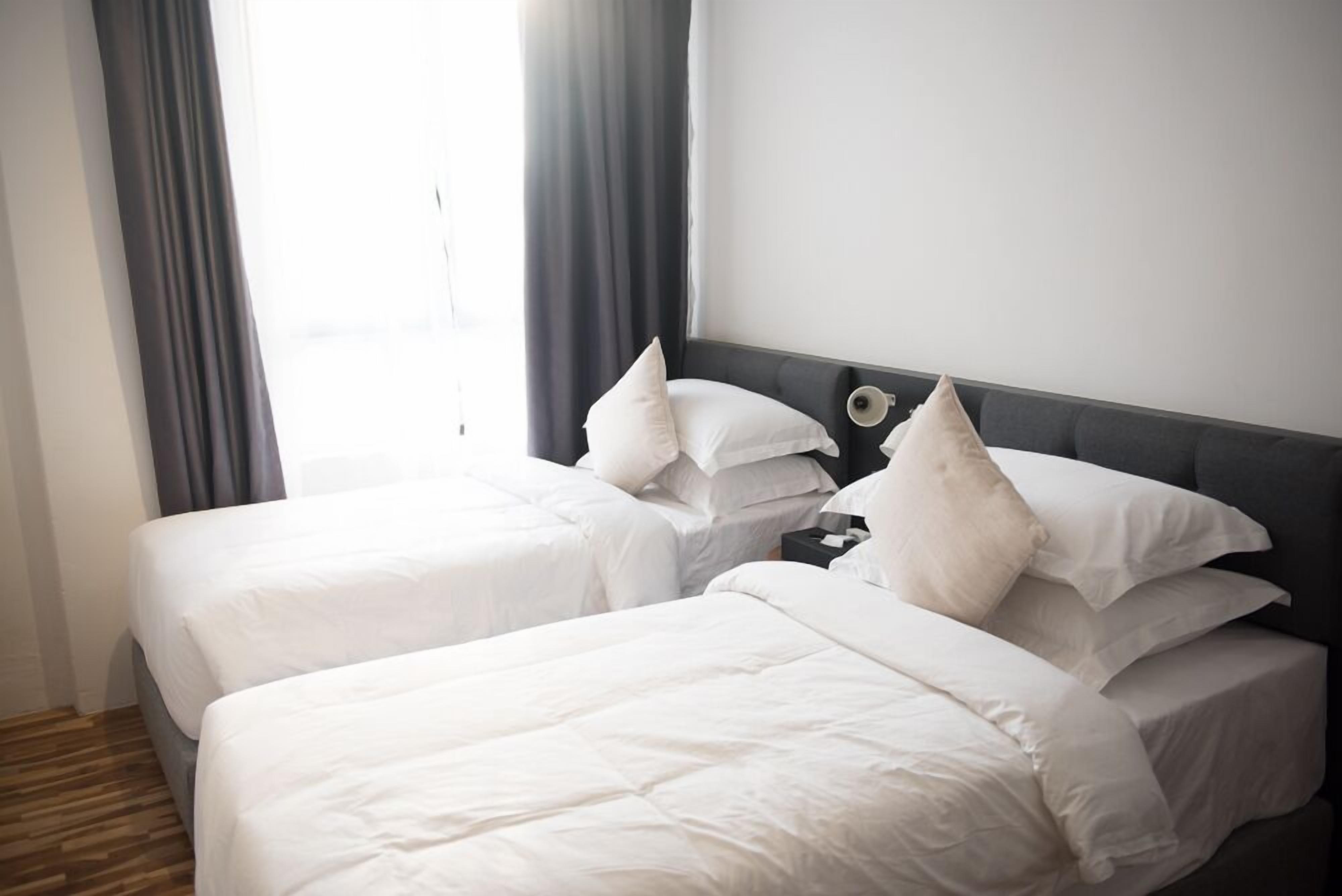 Une chambre d’hôtel avec deux lits, chacun doté de draps blancs et de plusieurs oreillers. Il y a une table de chevet avec une lampe et une petite table entre les lits. La chambre est équipée d’une grande fenêtre avec des rideaux, ce qui permet à la lumière naturelle d’entrer.