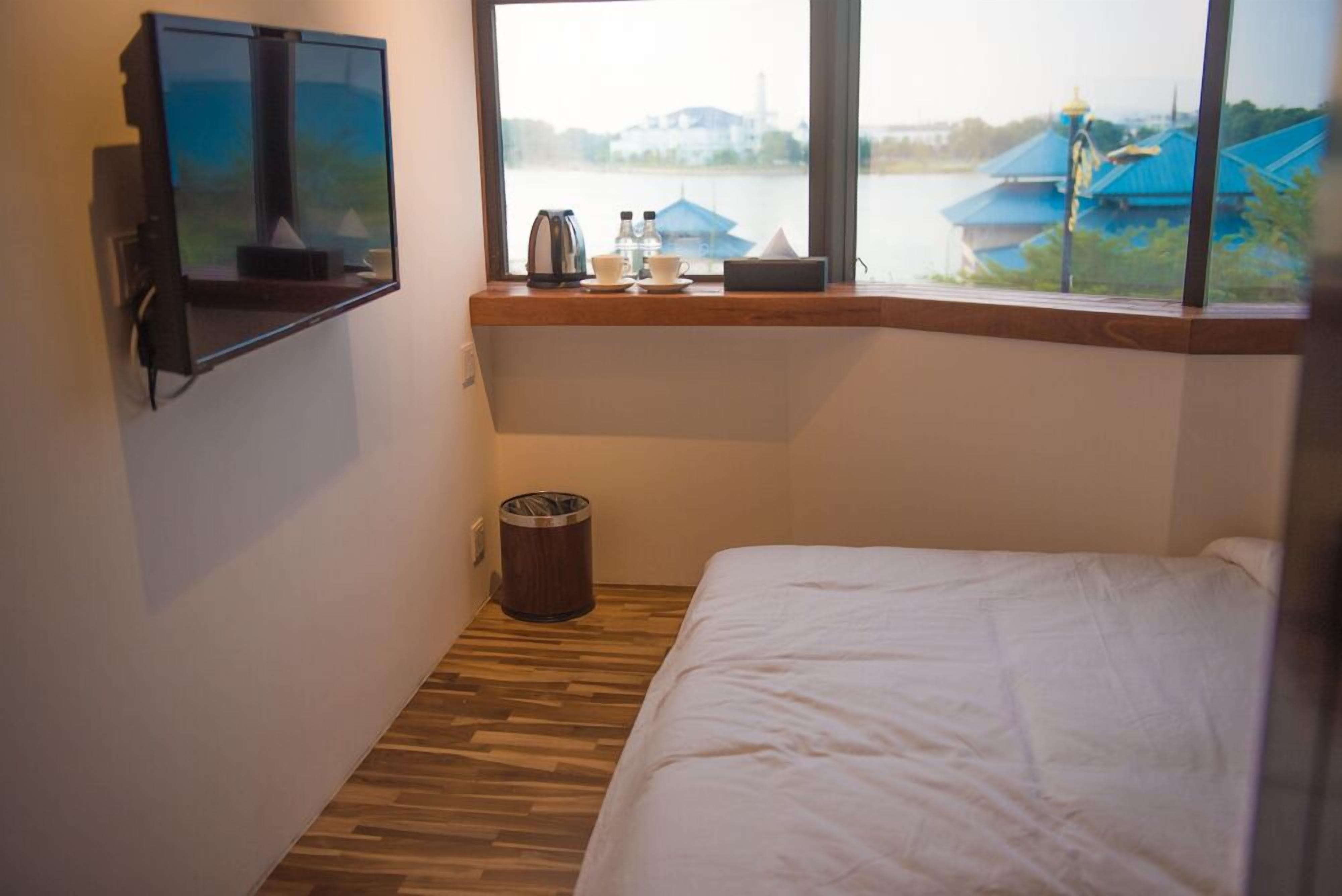 A small hotel room with a bed, a TV mounted on the wall, a window with a view of buildings and a body of water, and a trash bin.