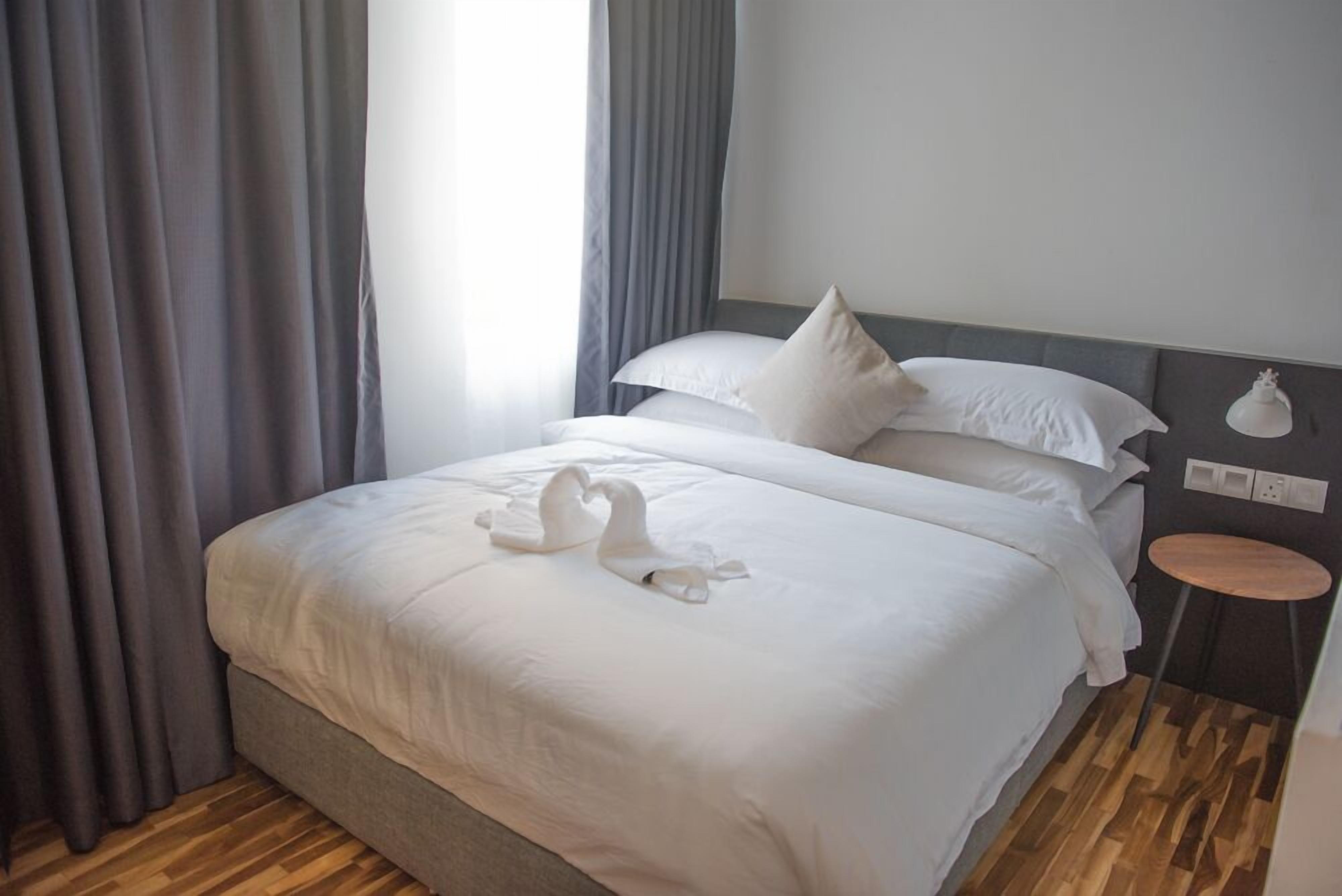 A neatly made bed with white linens and two swan-shaped pillows, a small round table beside the bed, and a grey curtain partially drawn to the side.