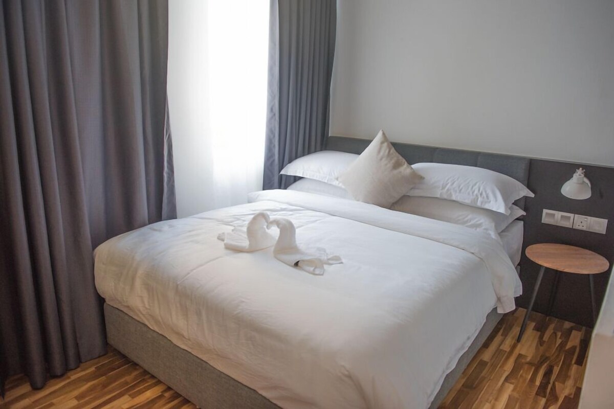 A neatly made bed with white linens and two swan-shaped pillows, a small round table beside the bed, and a grey curtain partially drawn to the side.