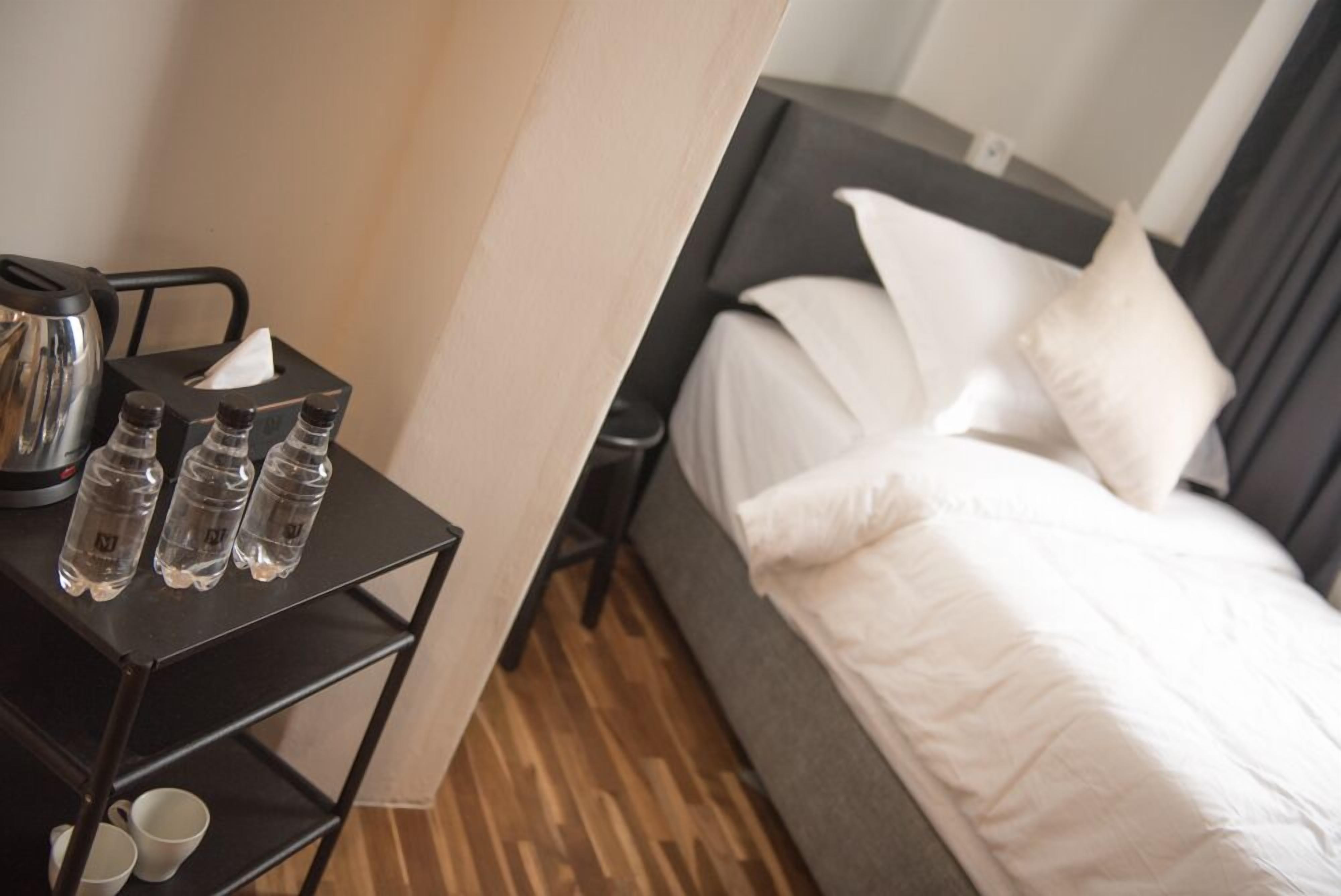 Quarto de hotel com cama, mesa de cabeceira com chaleira, garrafas de água e copos.