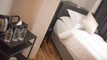 Quarto de hotel com cama, mesa de cabeceira com chaleira, garrafas de água e copos.