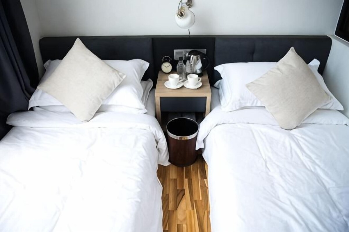 A hotel room with two beds, each with white bedding and beige pillows, a small wooden nightstand with a kettle, cups, and a clock, and a wooden floor.