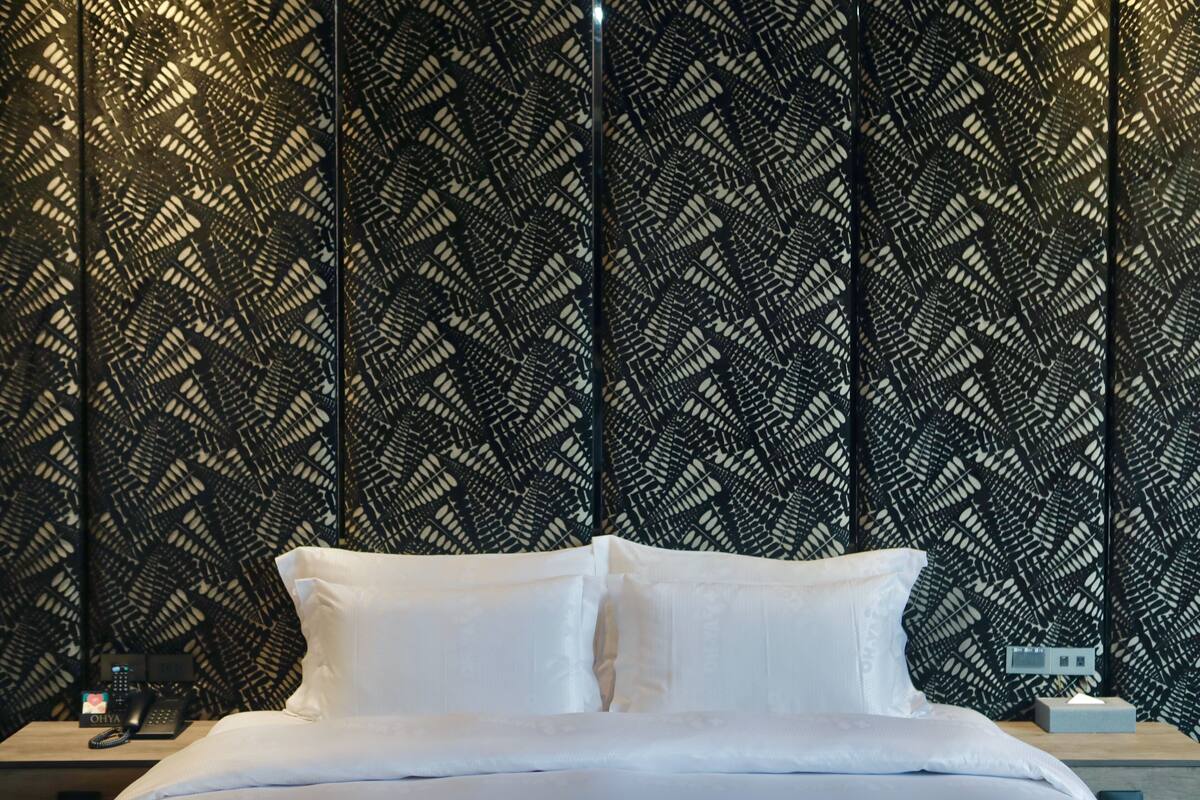 Un lit avec du linge de lit blanc, une table de chevet avec un téléphone et un mur orné d’un motif géométrique.