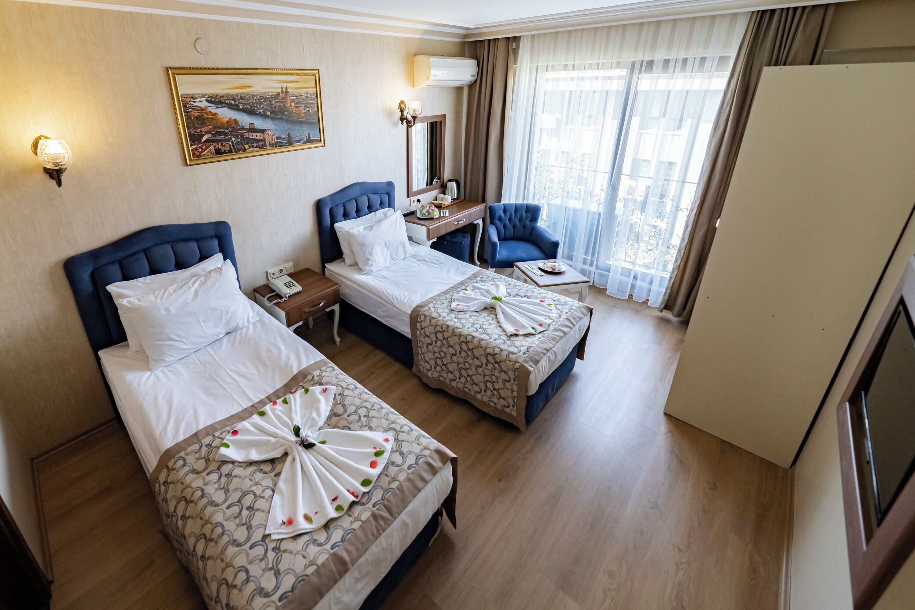 Une chambre d’hôtel avec deux lits, un bureau, une chaise, une grande fenêtre avec des rideaux et un tableau au mur.