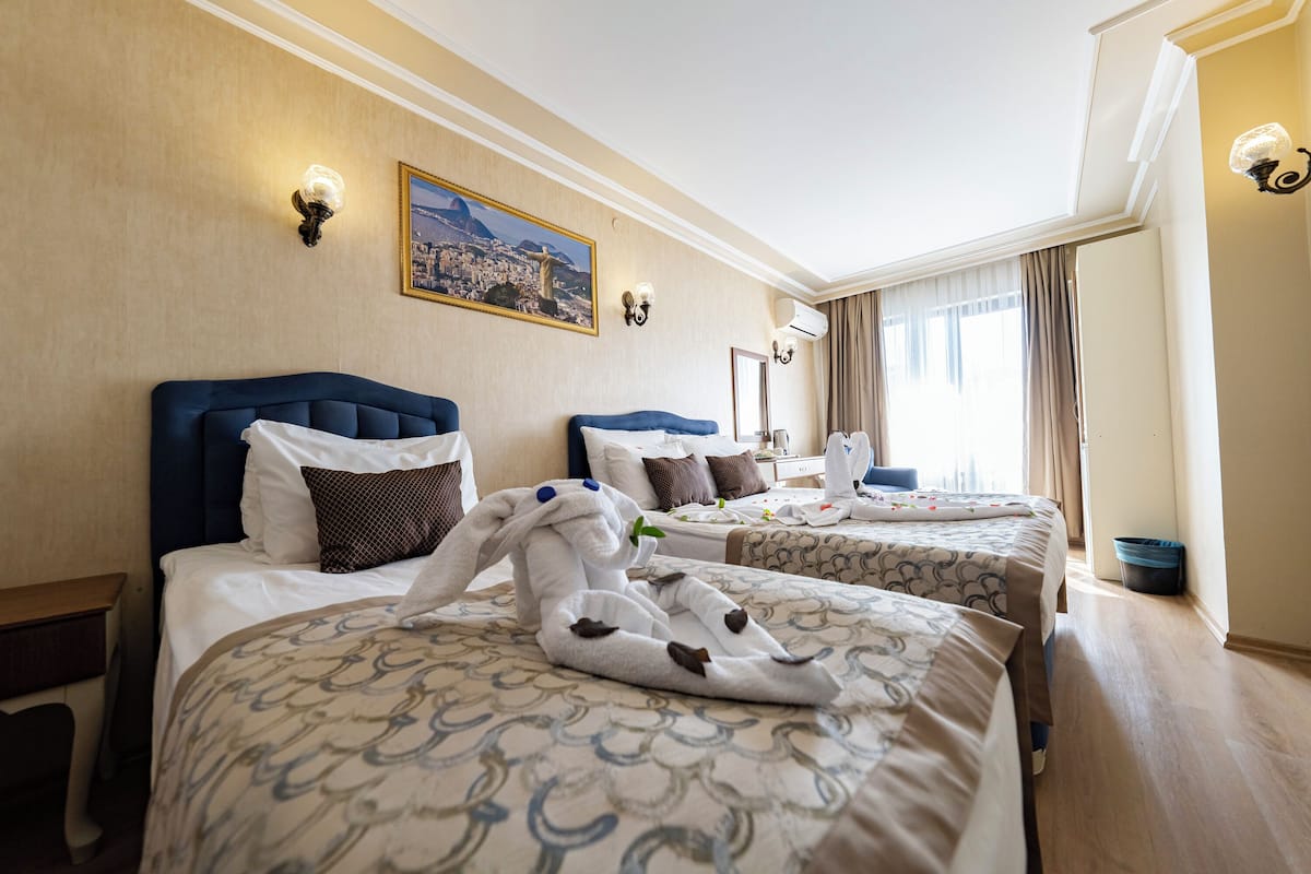 Habitació d'hotel amb dos llits, cadascun amb un capçal blau, roba de llit blanca i una tovallola amb forma de conill. Hi ha un quadre a la paret i una finestra amb cortines.
