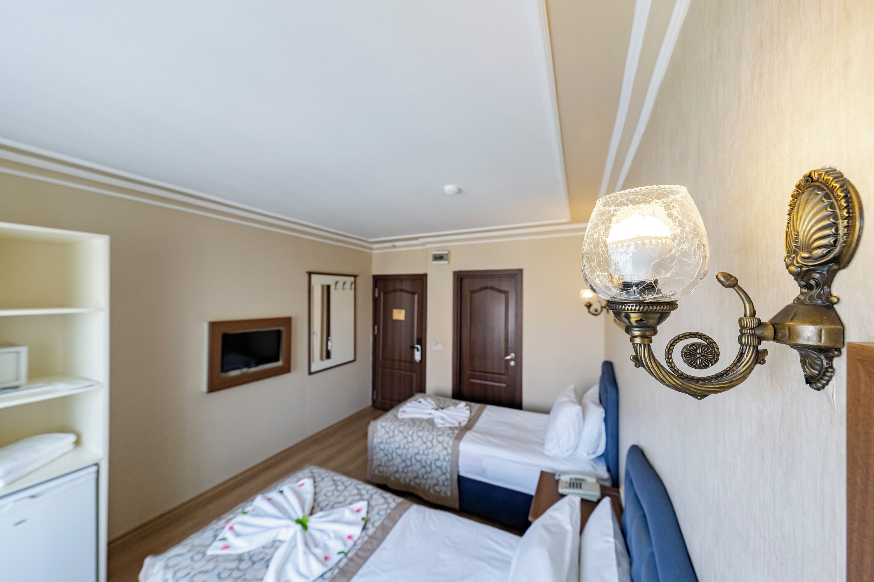 A hotel room with two beds, a wardrobe, a desk, and a wall-mounted light fixture.