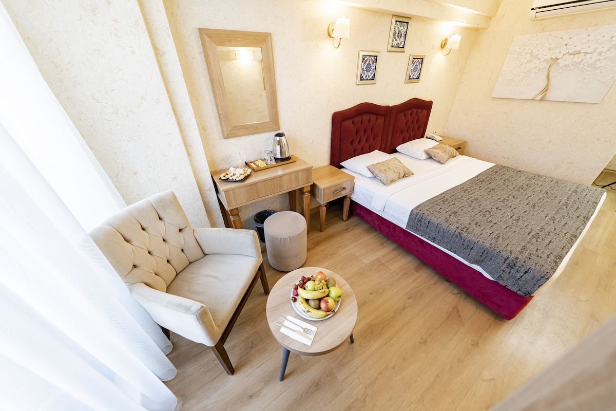 A hotel room with a bed, a desk, a chair, a small table with fruit, and a trash can.