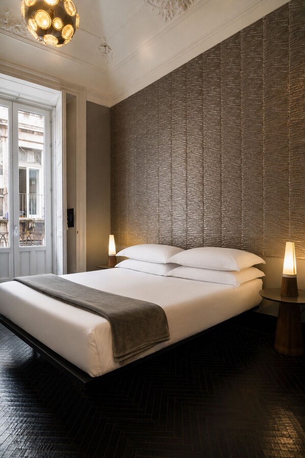 Una habitación de hotel moderna con una cama grande, un muro con textura, piso de madera y un candelabro.