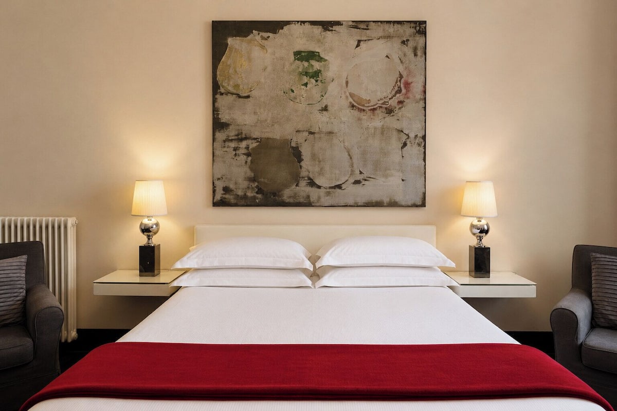 Ein Hotelzimmer mit einem Bett, zwei Nachttischen mit Lampen, einem Heizkörper und einem großen abstrakten Gemälde an der Wand.