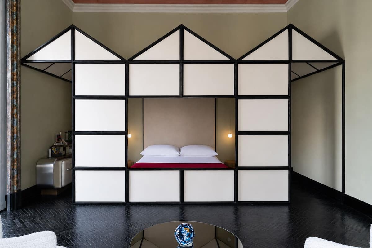 Ein Zimmer mit einem Bett in der Mitte, einem kleinen Tisch mit einem dekorativen Gegenstand und einer Wand mit geometrischem Muster.