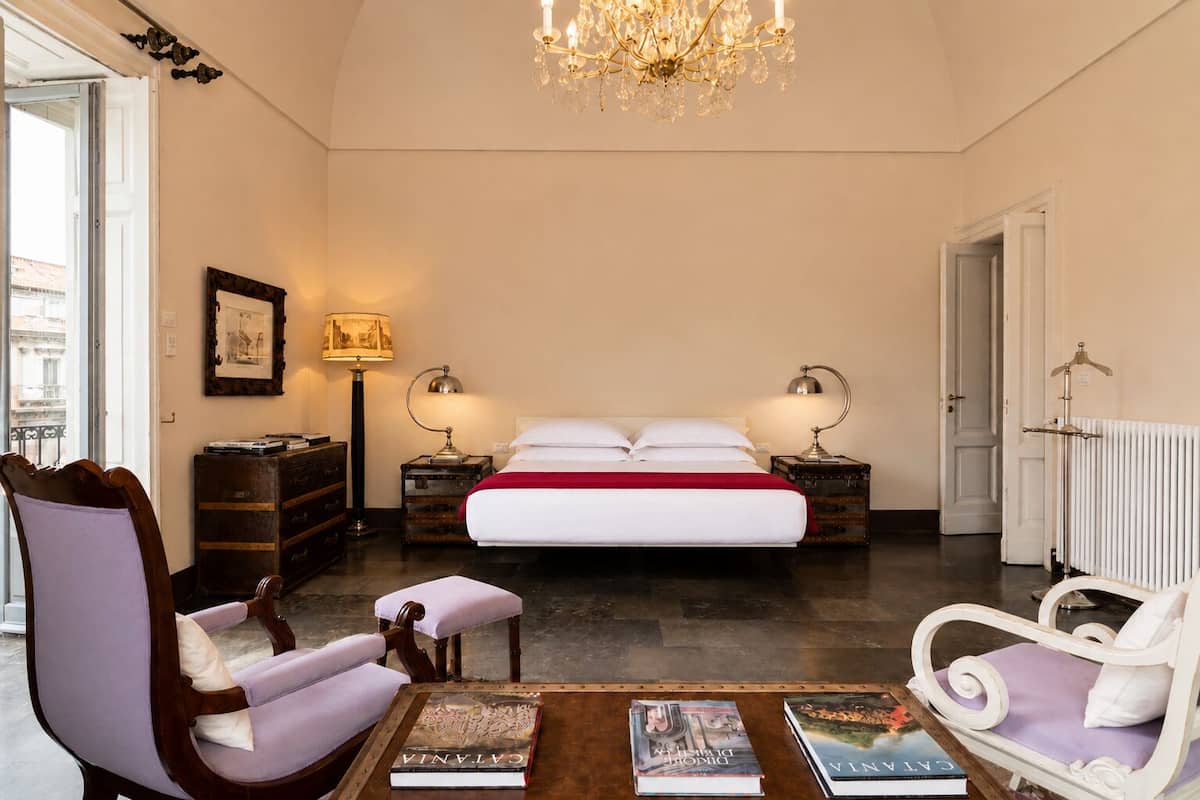 Ein Schlafzimmer mit einem großen Bett, zwei Nachttischlampen, einem Kronleuchter, einem Schminktisch, einem Sessel und einem Tisch mit Büchern.