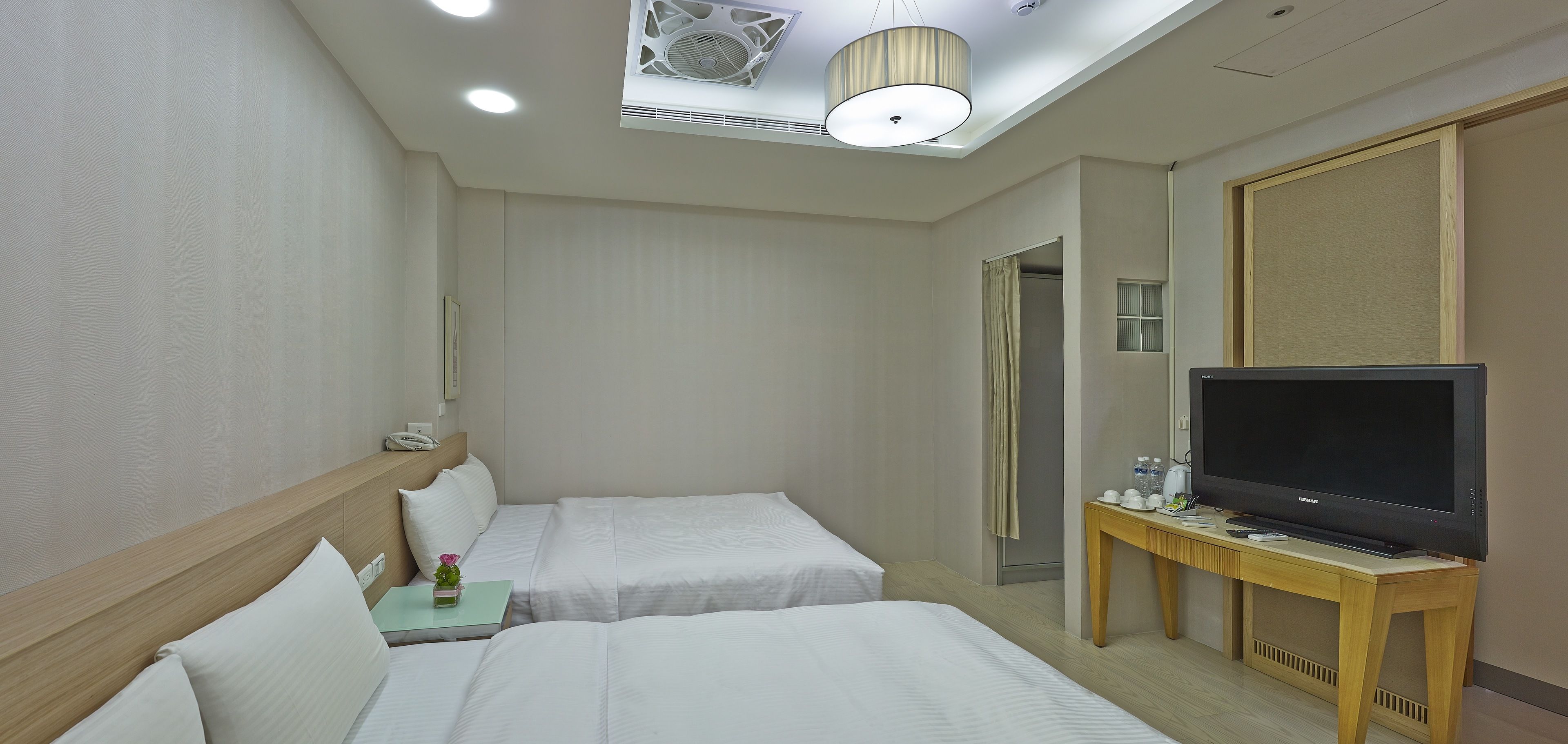 A hotel room with two beds, a TV on a wooden stand, a small table with a phone and a flower vase, and a ceiling fan.