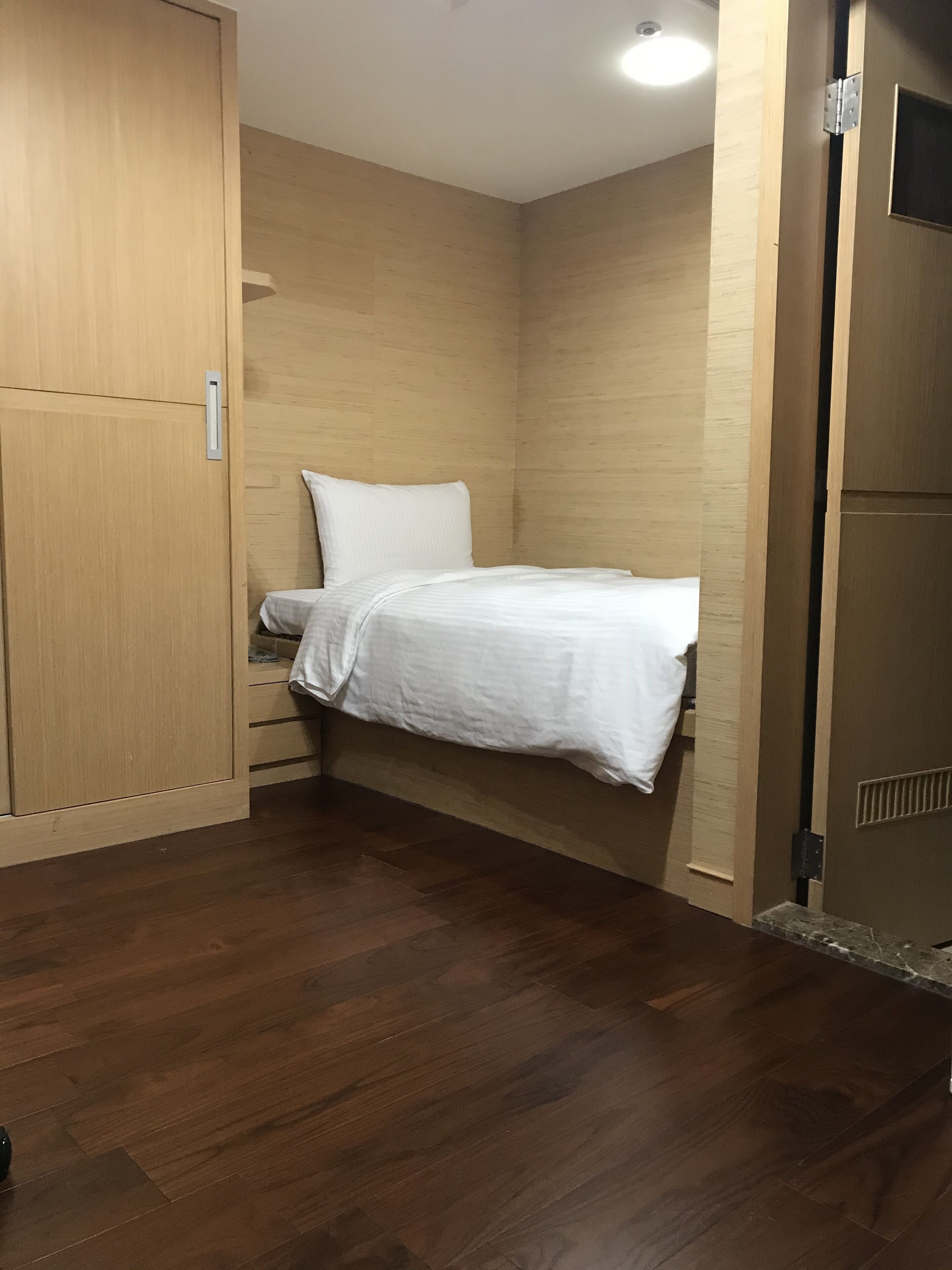 A small, compact room with a bed, wooden wardrobe, and a door.