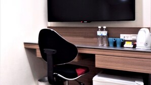A hotel room with a desk, a black office chair, a flat-screen TV mounted on the wall, a small refrigerator, and a trash bin. - West 59 Inn  (Taipei)