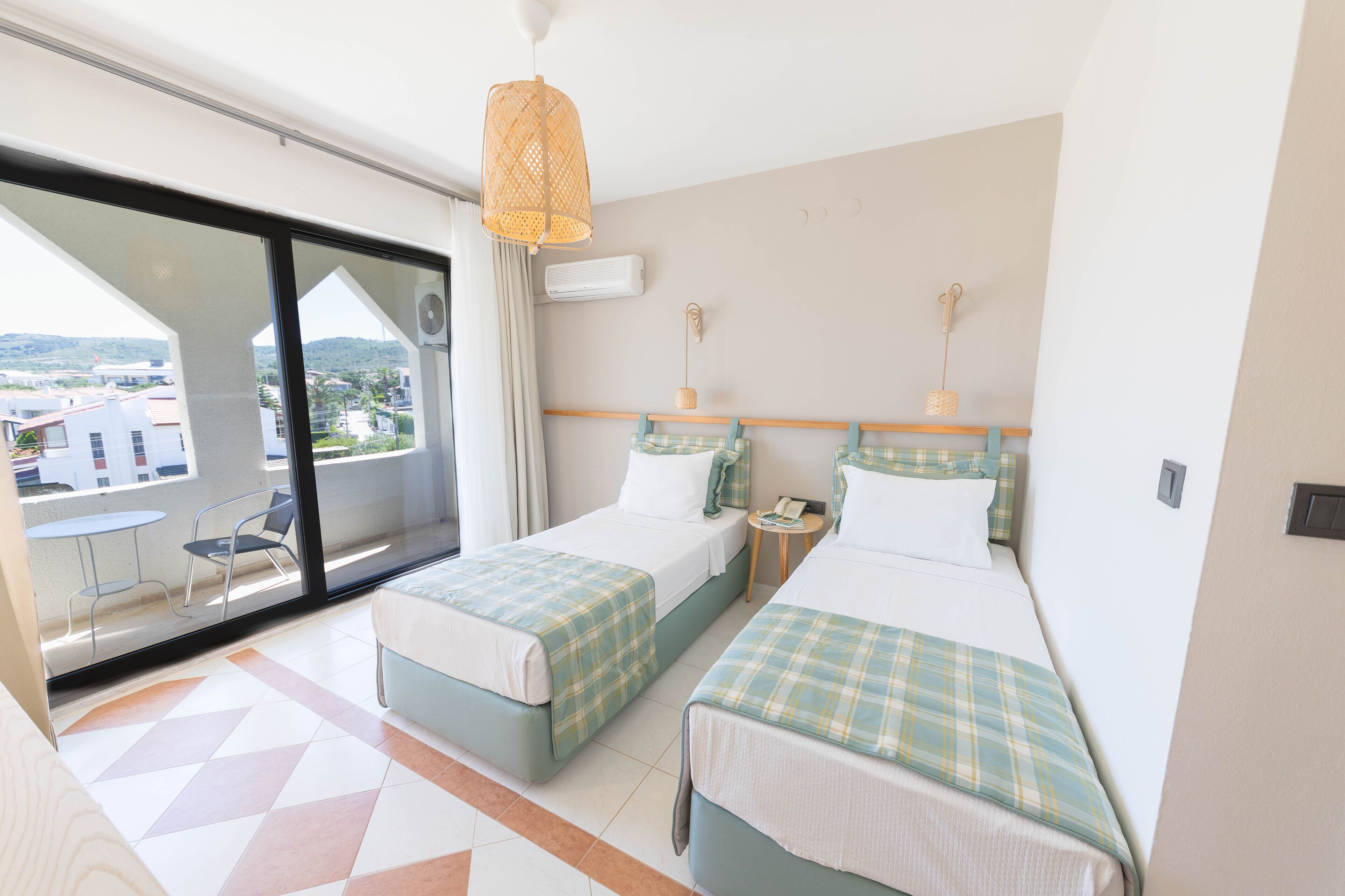 A hotel room with two beds, a balcony with a table and chairs, and a large window offering a city view.