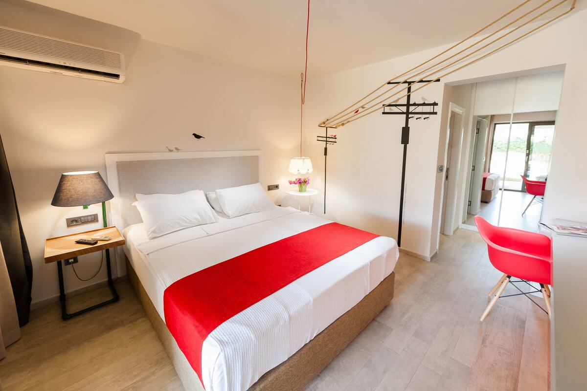 A modern hotel room with a large bed, bedside table, lamp, and a red chair.