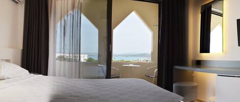 A hotel room with a bed, a desk, a chair, a small table, a trash bin, and a view of the city through a large window.