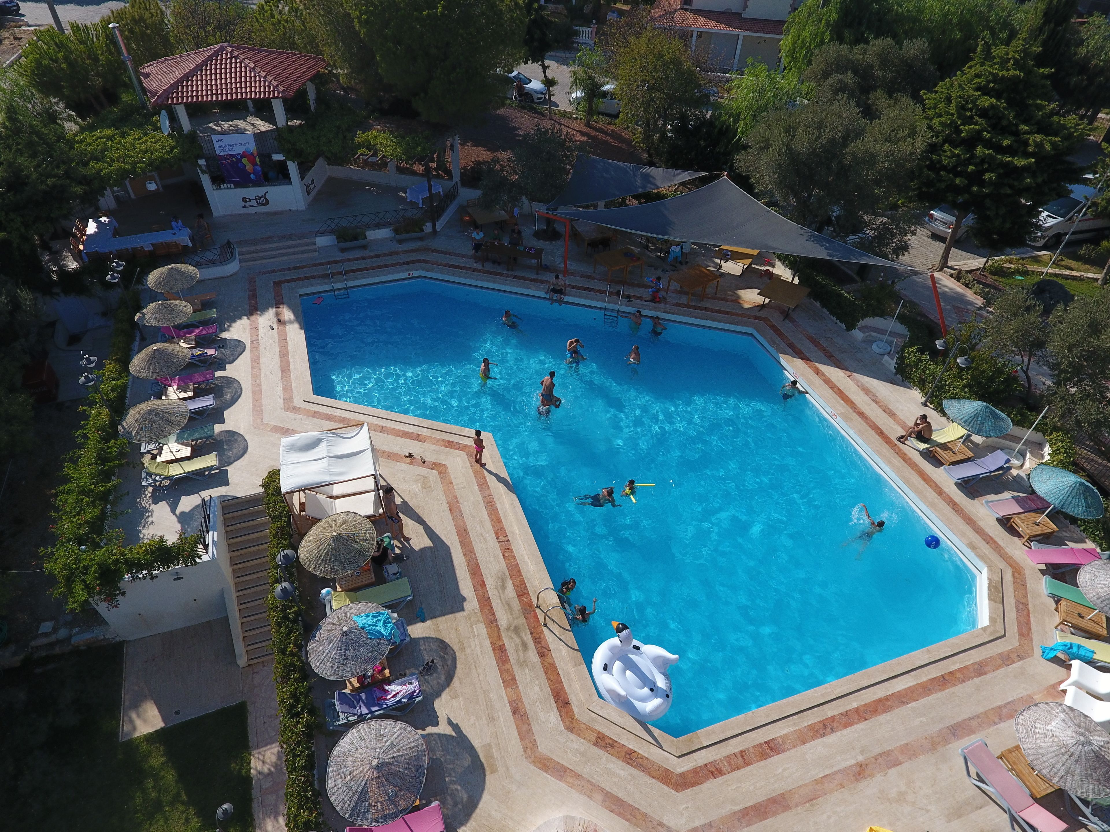 A large swimming pool surrounded by sun loungers and umbrellas, with a few people swimming and others relaxing on the deck.