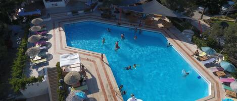 A large swimming pool surrounded by sun loungers and umbrellas, with a few people swimming and others relaxing on the deck.