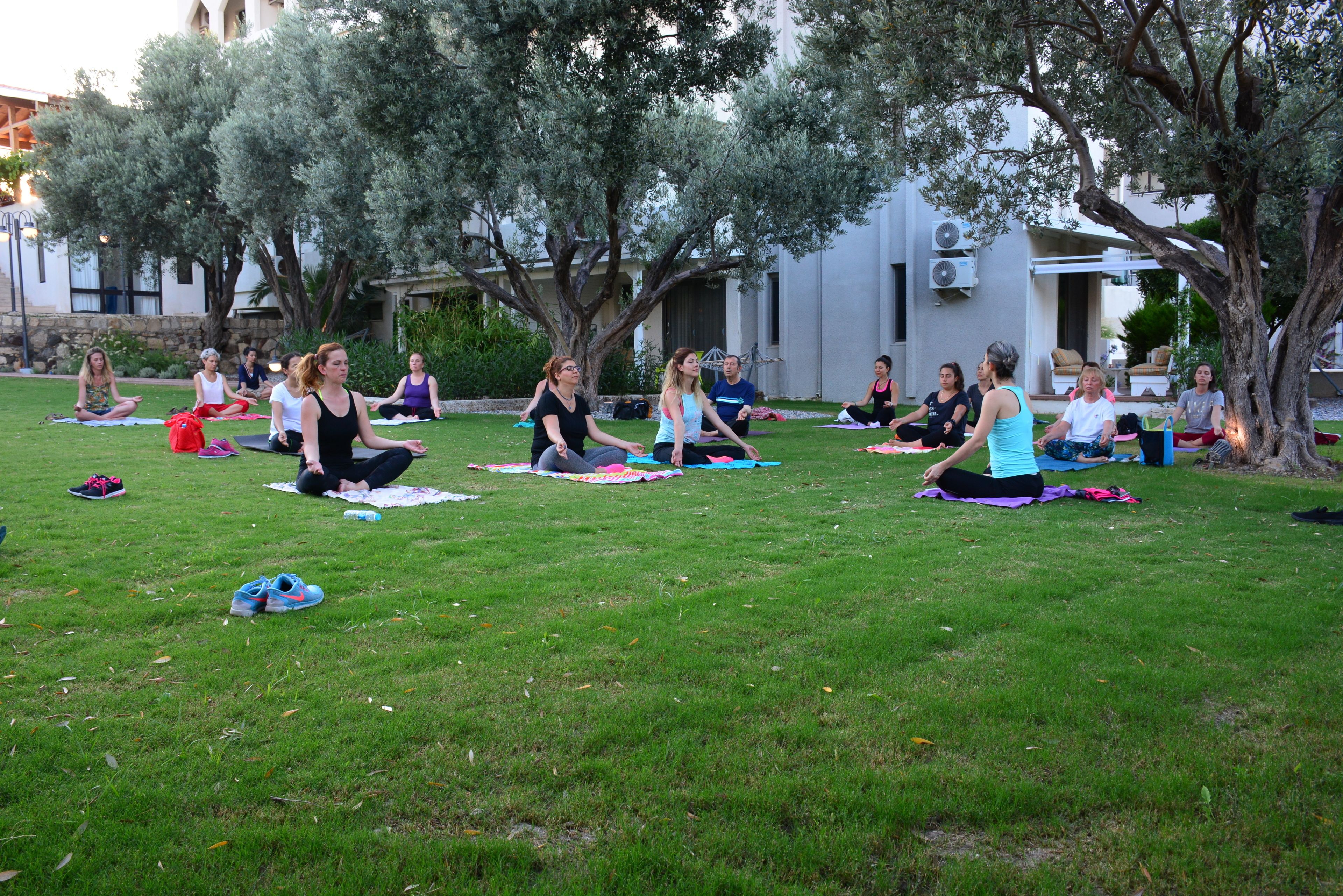 A group of people sitting on mats in a grassy outdoor area with trees and a building in the background.