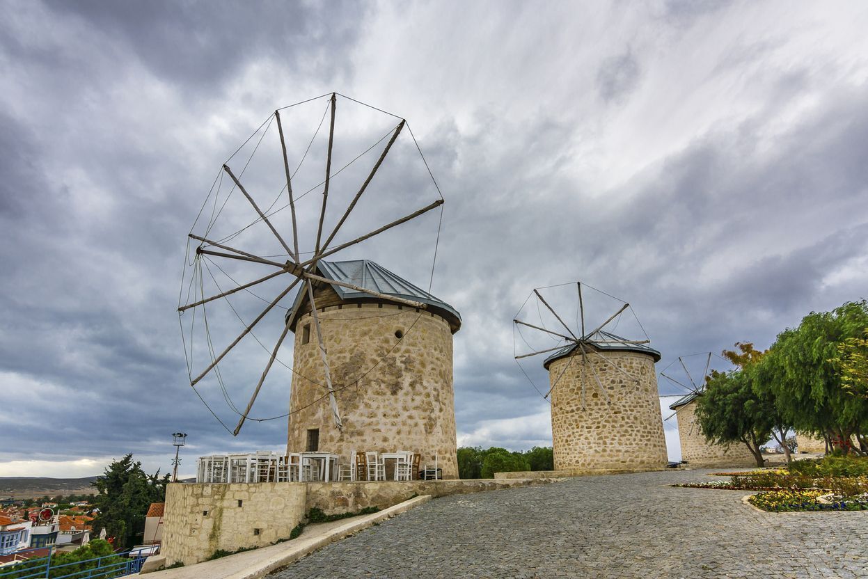 Two windmills with stone bases and metal blades, set on a cobblestone path with a view of a town in the background.
