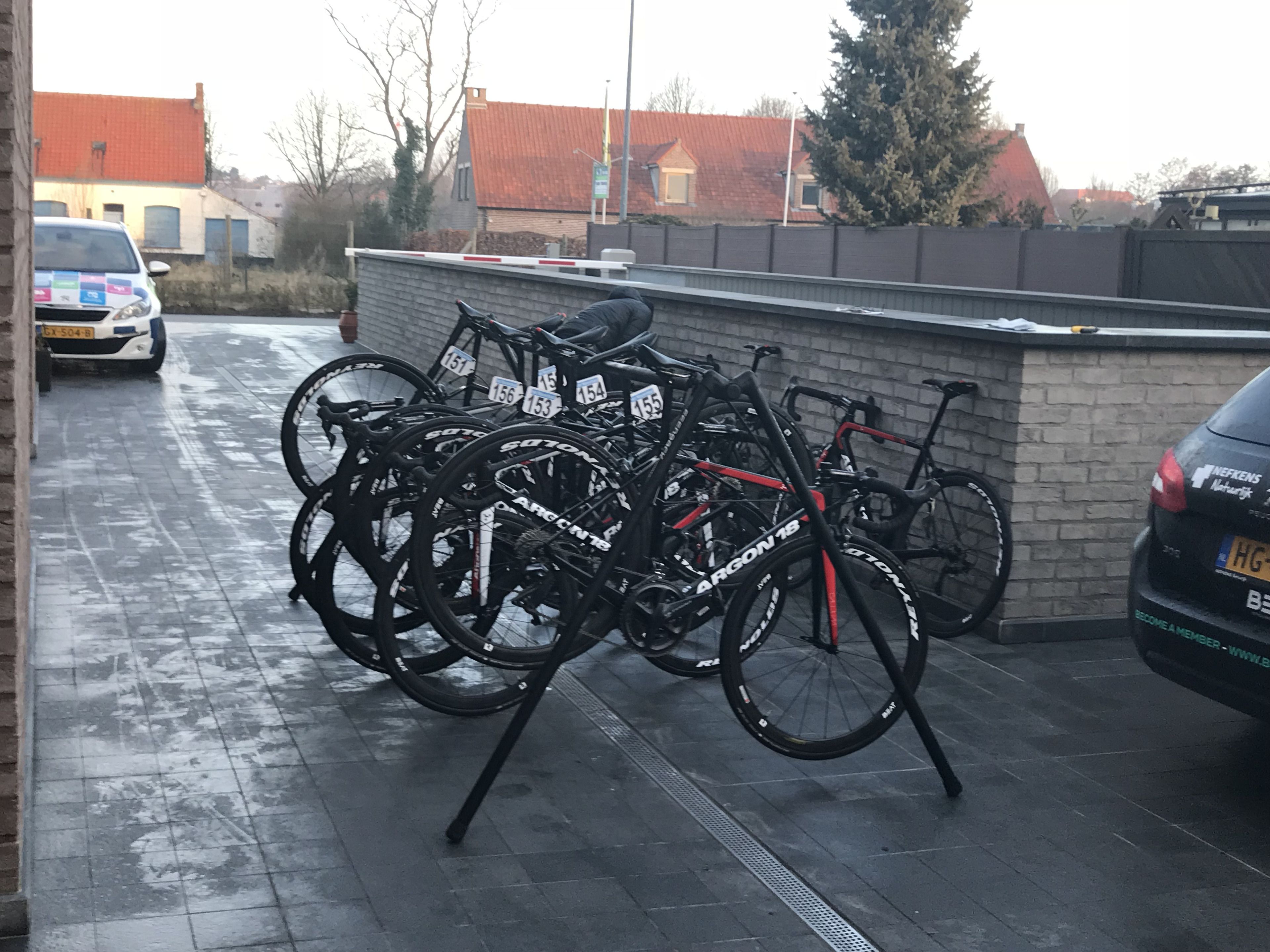 Bicycles parked in a covered area with a brick wall and a car in the background.