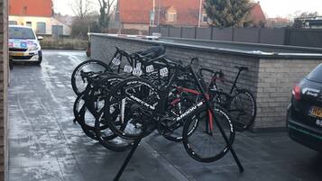 Bicycles parked in a covered area with a brick wall and a car in the background.