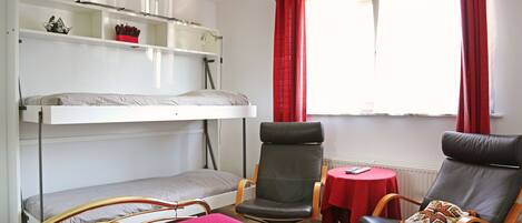 A room with a bunk bed, two chairs, a small table, and a window with curtains.