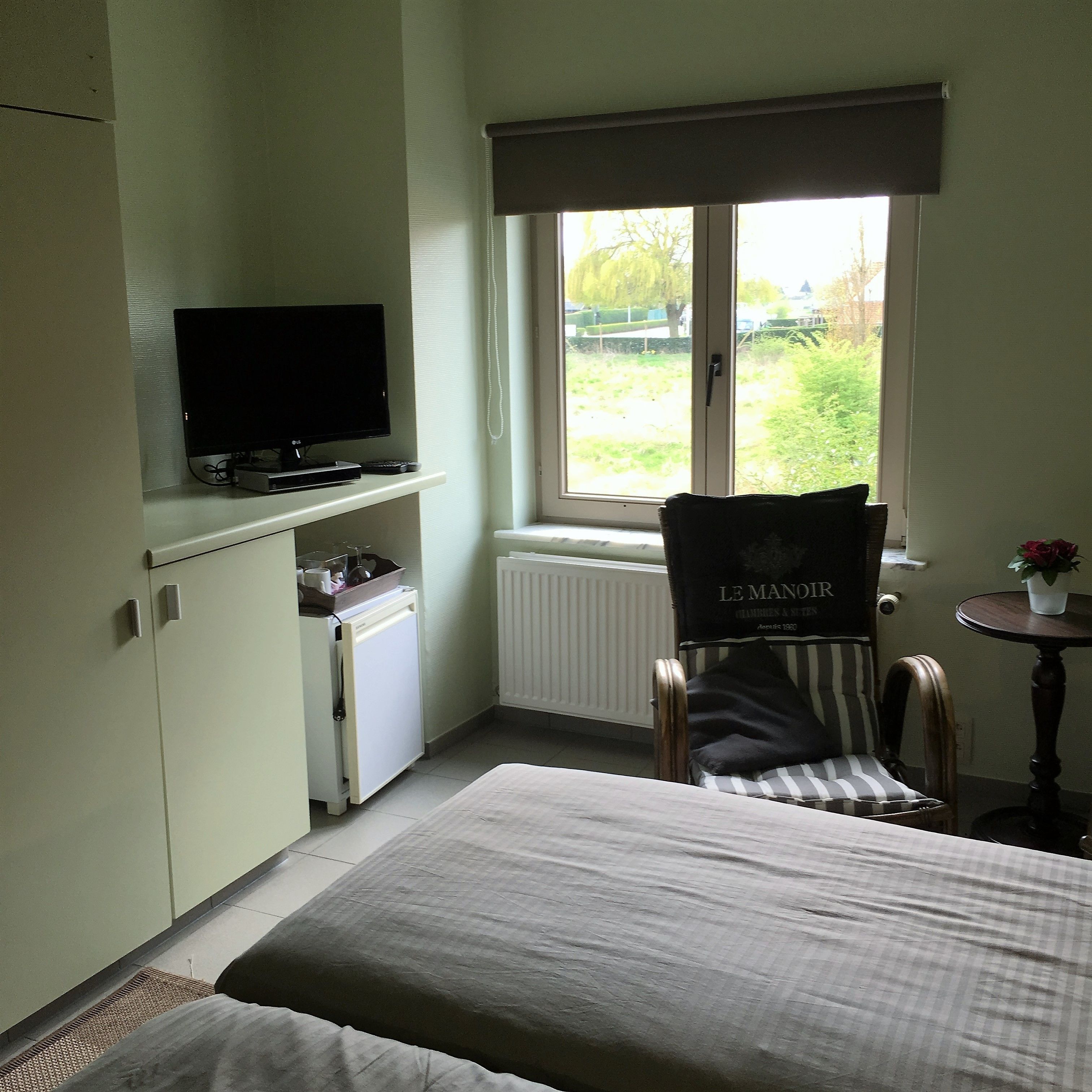 Una habitación pequeña y ordenada con una cama, una silla, un televisor, una mesita y una ventana con vistas a zonas verdes.
