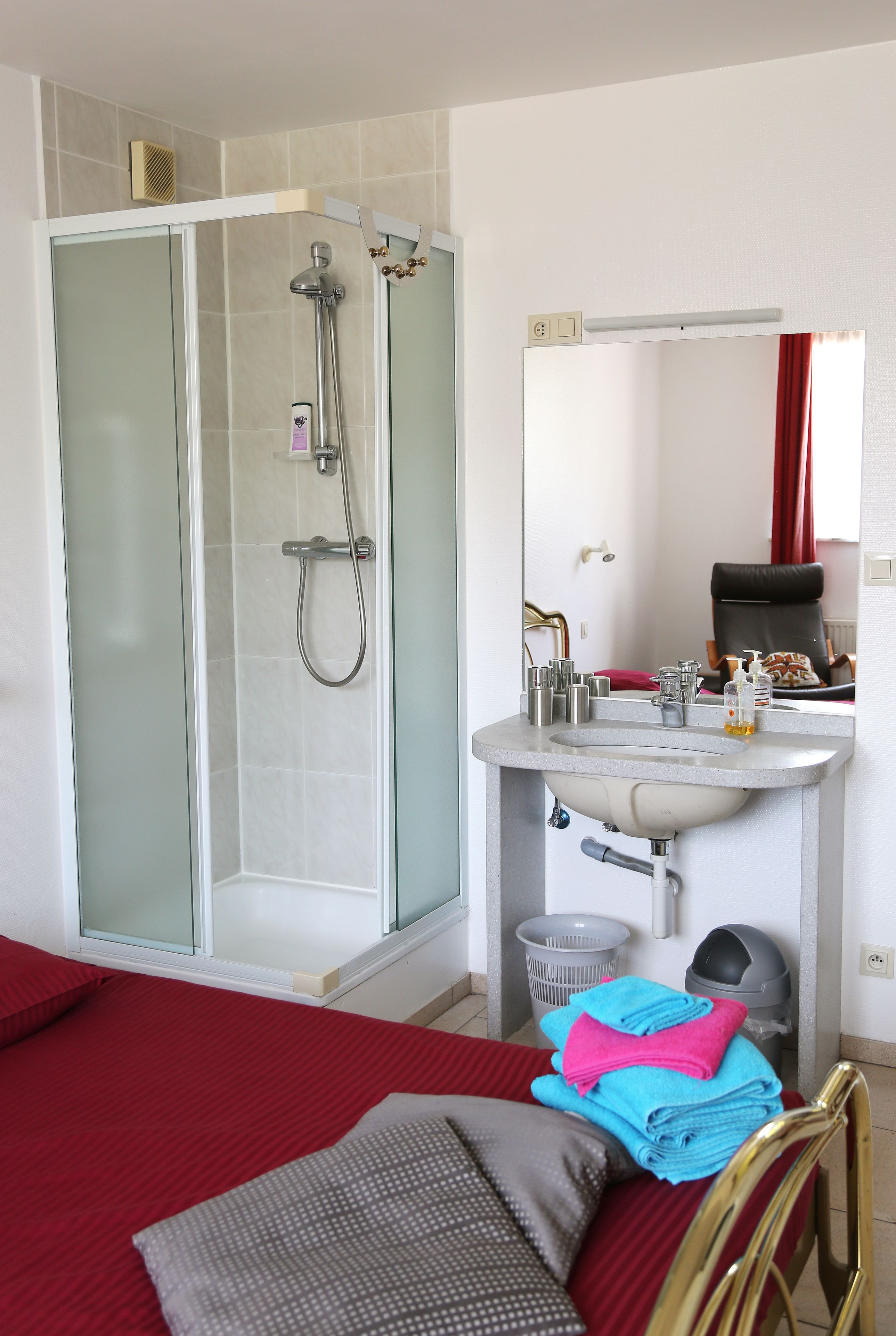 A hotel bathroom with a glass-enclosed shower, a sink with toiletries, and a bed with red bedding.