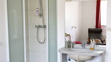 A hotel bathroom with a glass-enclosed shower, a sink with toiletries, and a bed with red bedding.