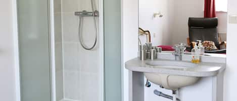 A hotel bathroom with a glass-enclosed shower, a sink with toiletries, and a bed with red bedding.