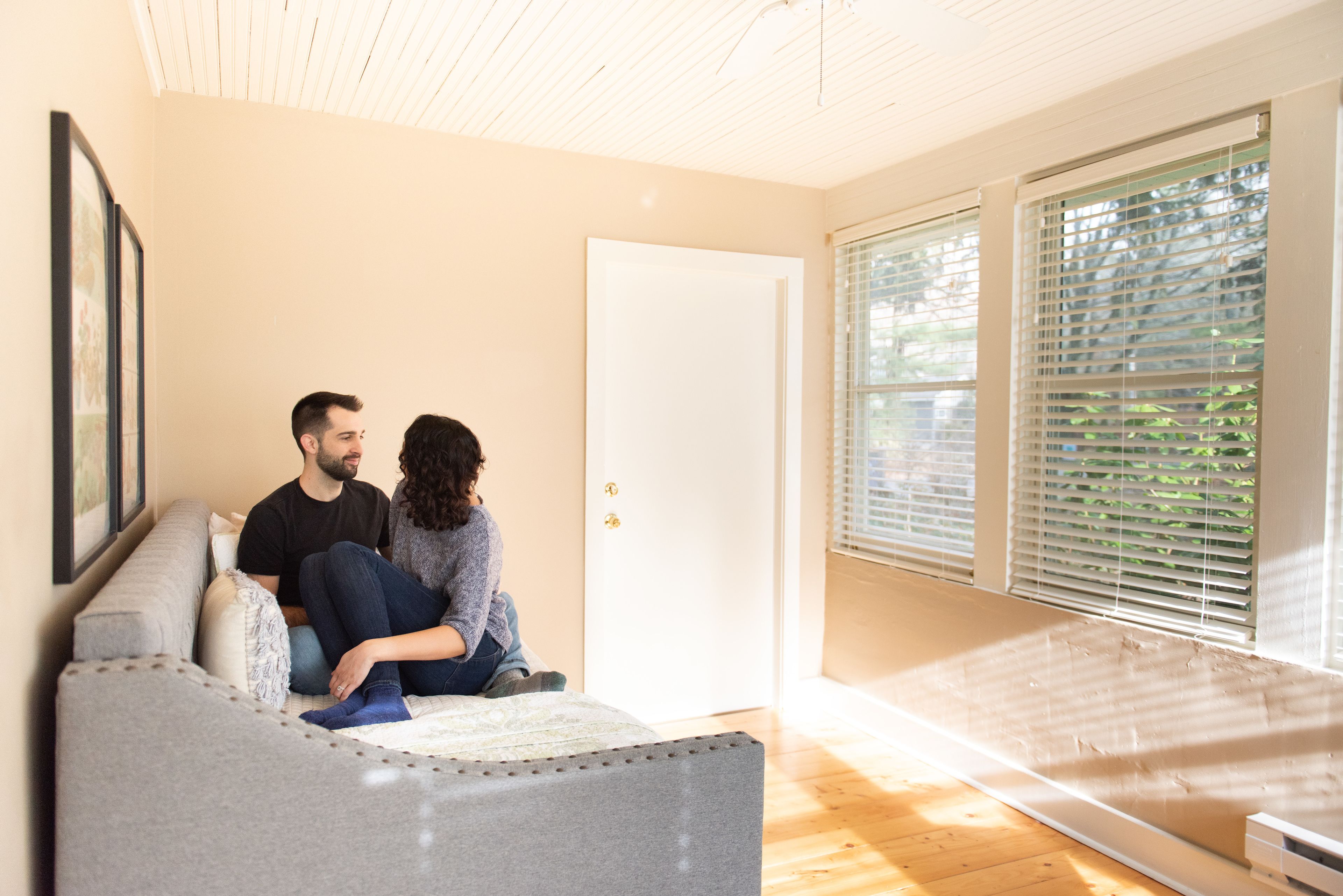 Una pareja sentada en un sofá gris en una habitación con una puerta blanca y ventanas amplias con persianas.