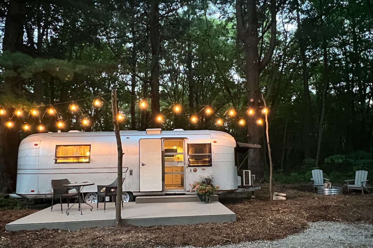 A white RV with a kitchen area, a dining table, and chairs, surrounded by trees and string lights.