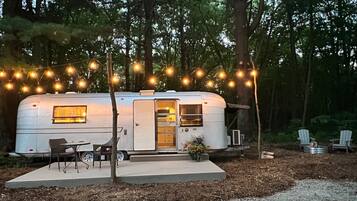 A white RV with a kitchen area, a dining table, and chairs, surrounded by trees and string lights.