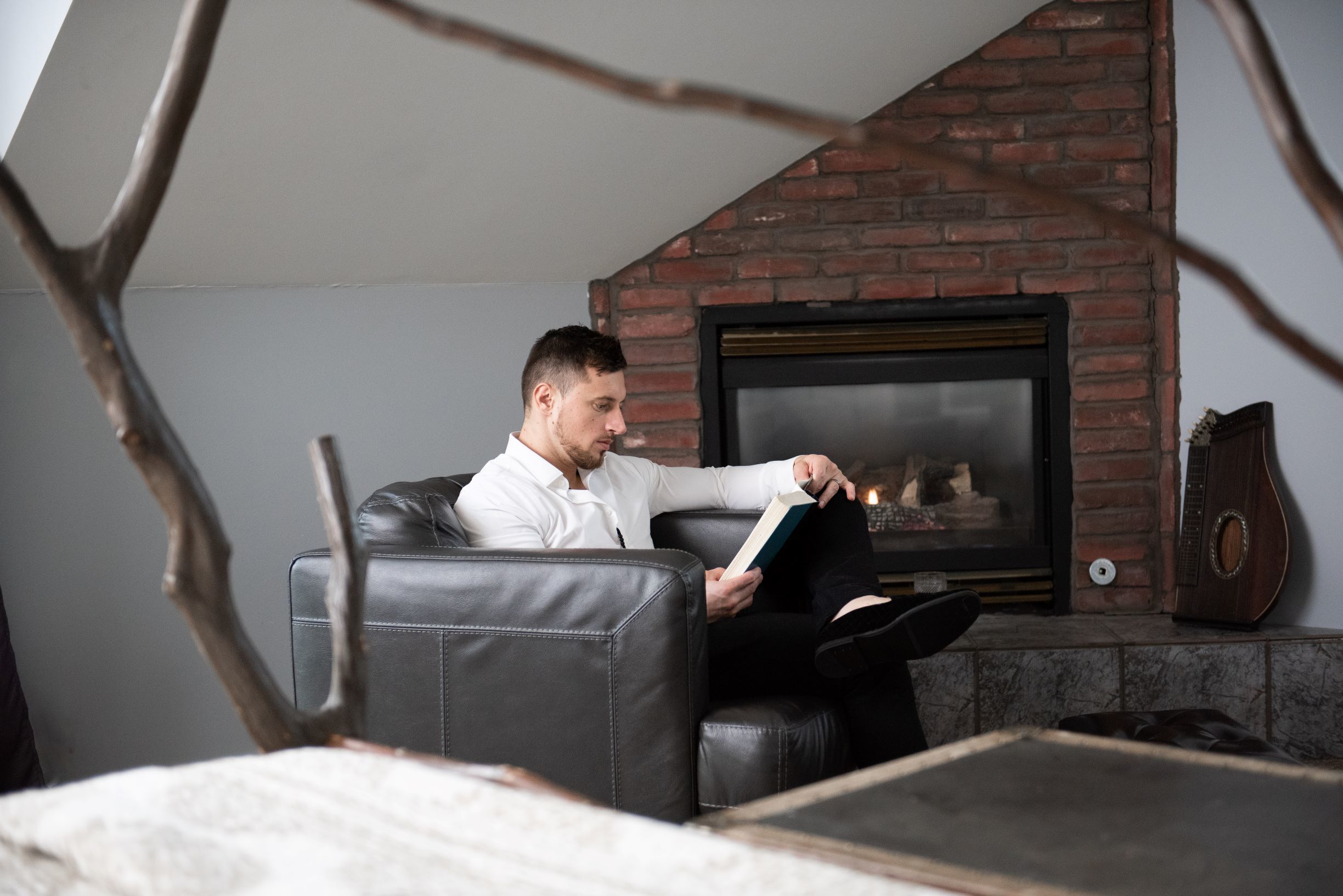 A man sitting on a leather couch reading a book in a room with a brick fireplace.