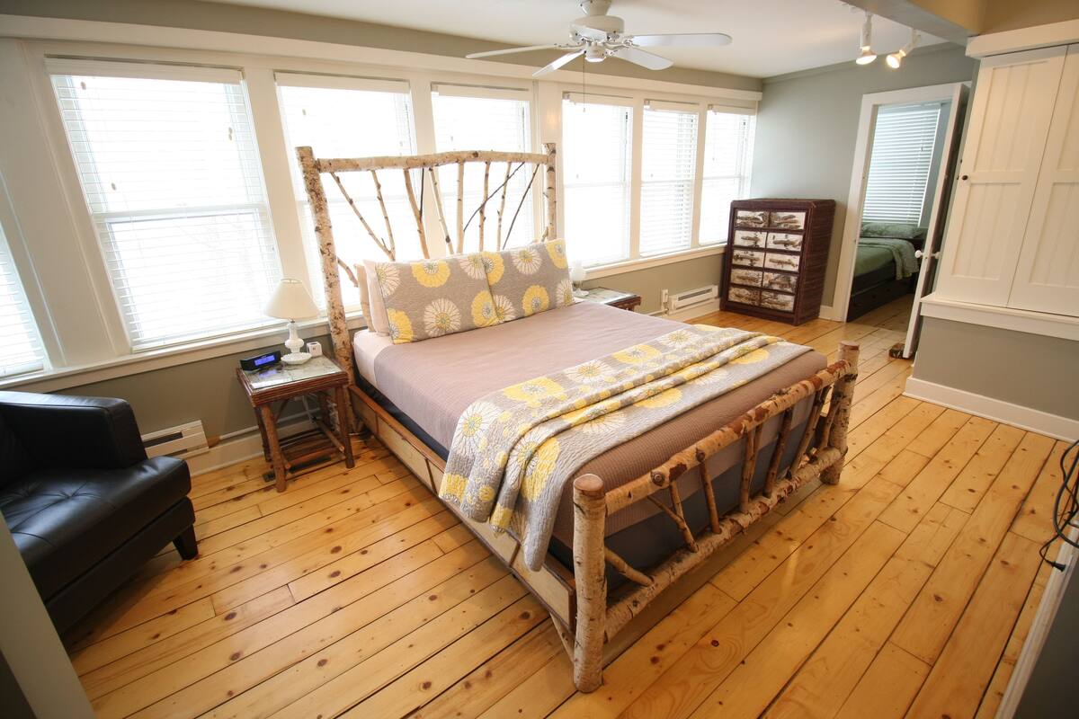 A bedroom with a wooden bed, a nightstand, a lamp, a dresser, and a window with blinds.