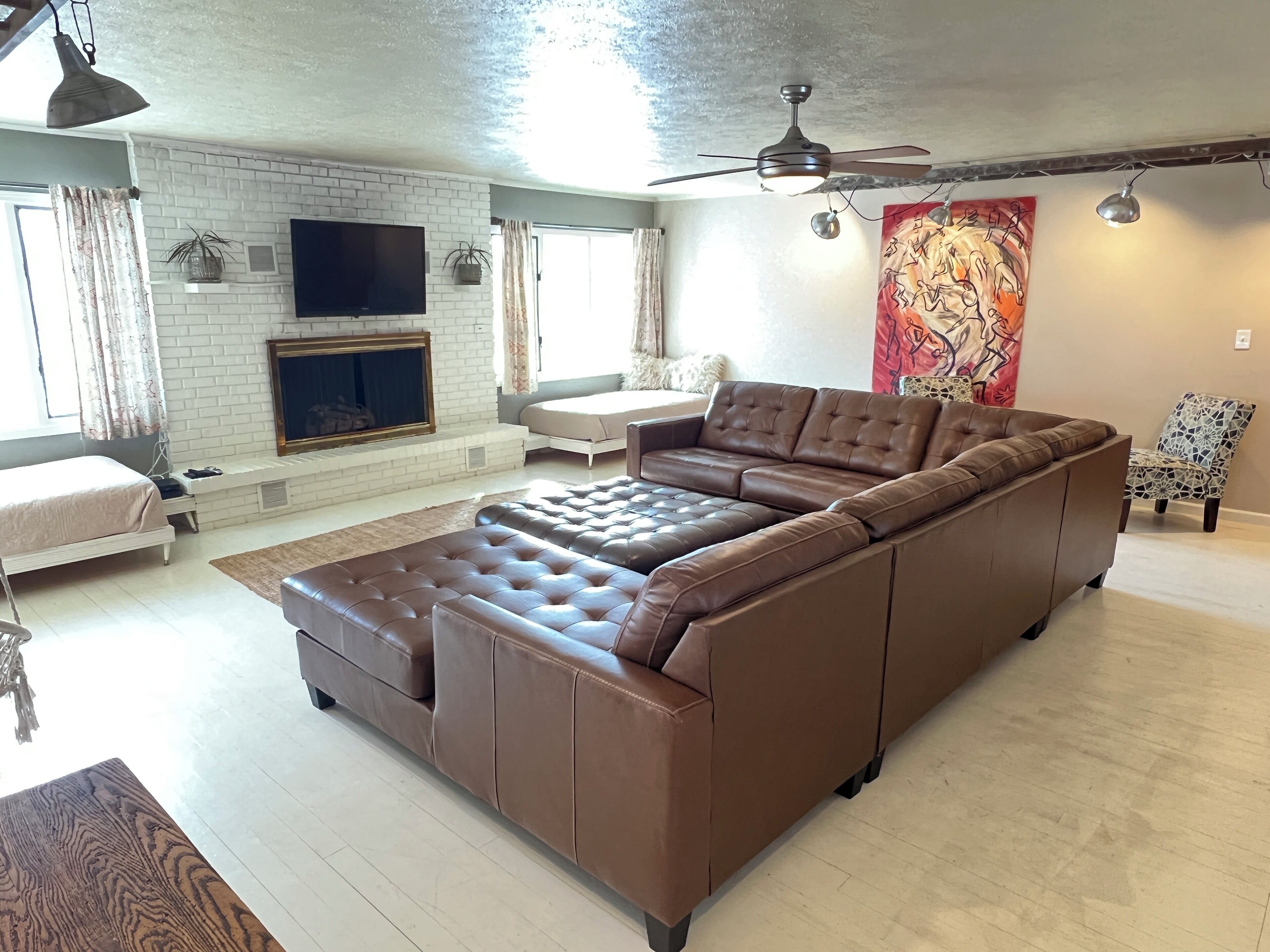 A spacious living room with a large brown sectional sofa, a flat-screen TV mounted on a white brick wall, a ceiling fan, and a red abstract painting.