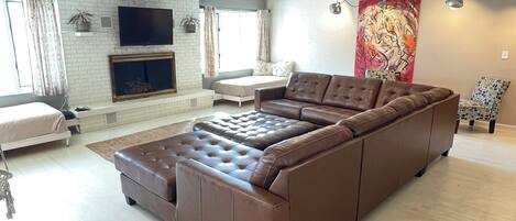 A spacious living room with a large brown sectional sofa, a flat-screen TV mounted on a white brick wall, a ceiling fan, and a red abstract painting.