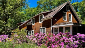 A two-story wooden house with a gabled roof, surrounded by a garden with vibrant pink flowers. - Goldberry Woods (Union Pier)