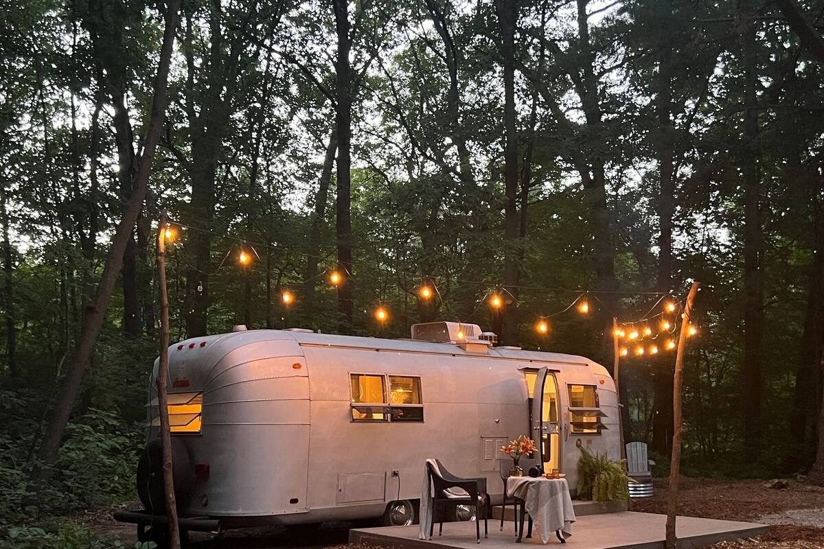 A stationary RV with a table and chairs outside, surrounded by trees and string lights.