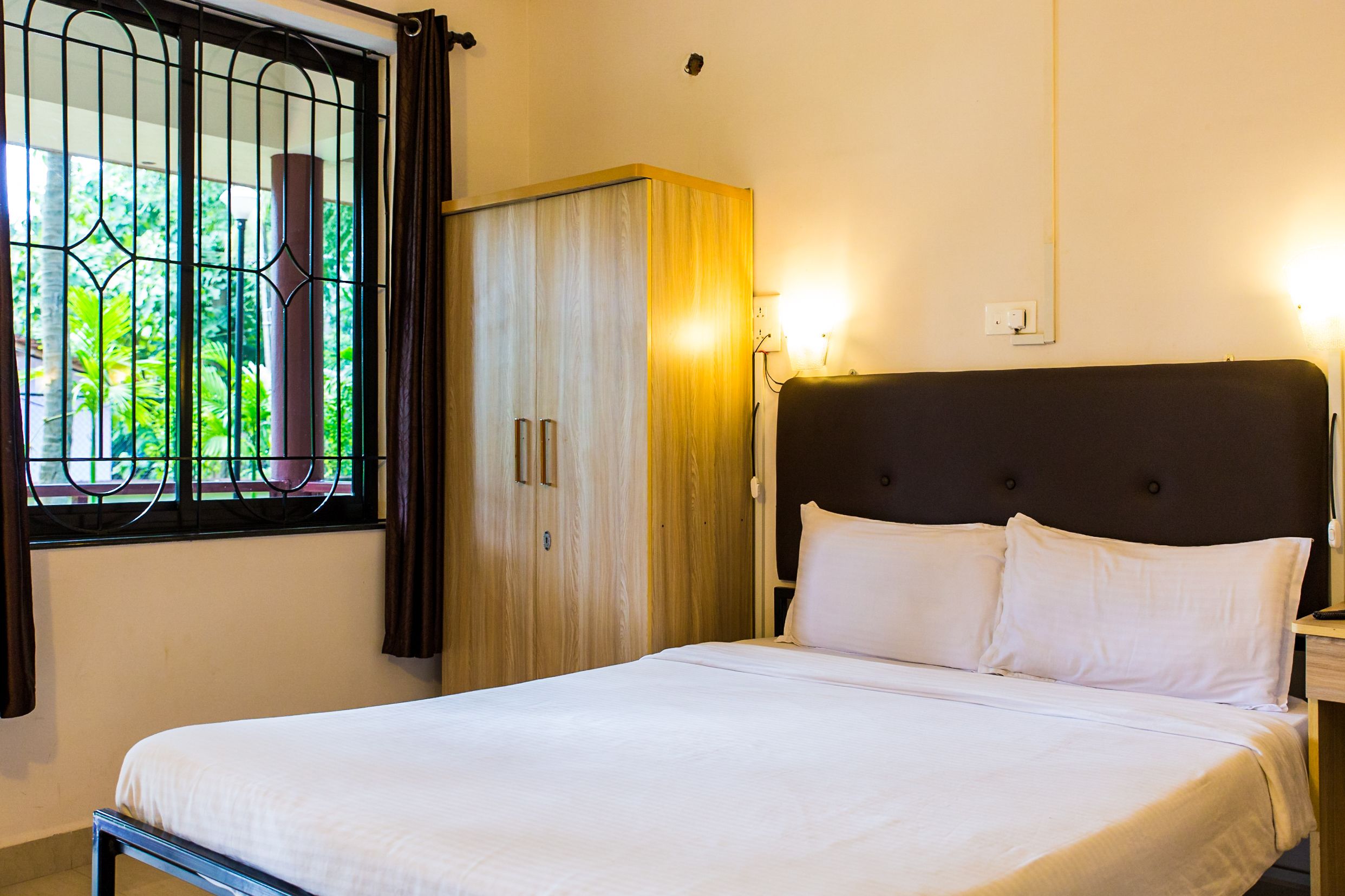 Una cama bien hecha con ropa blanca, un armario de madera y una ventana con barrotes.