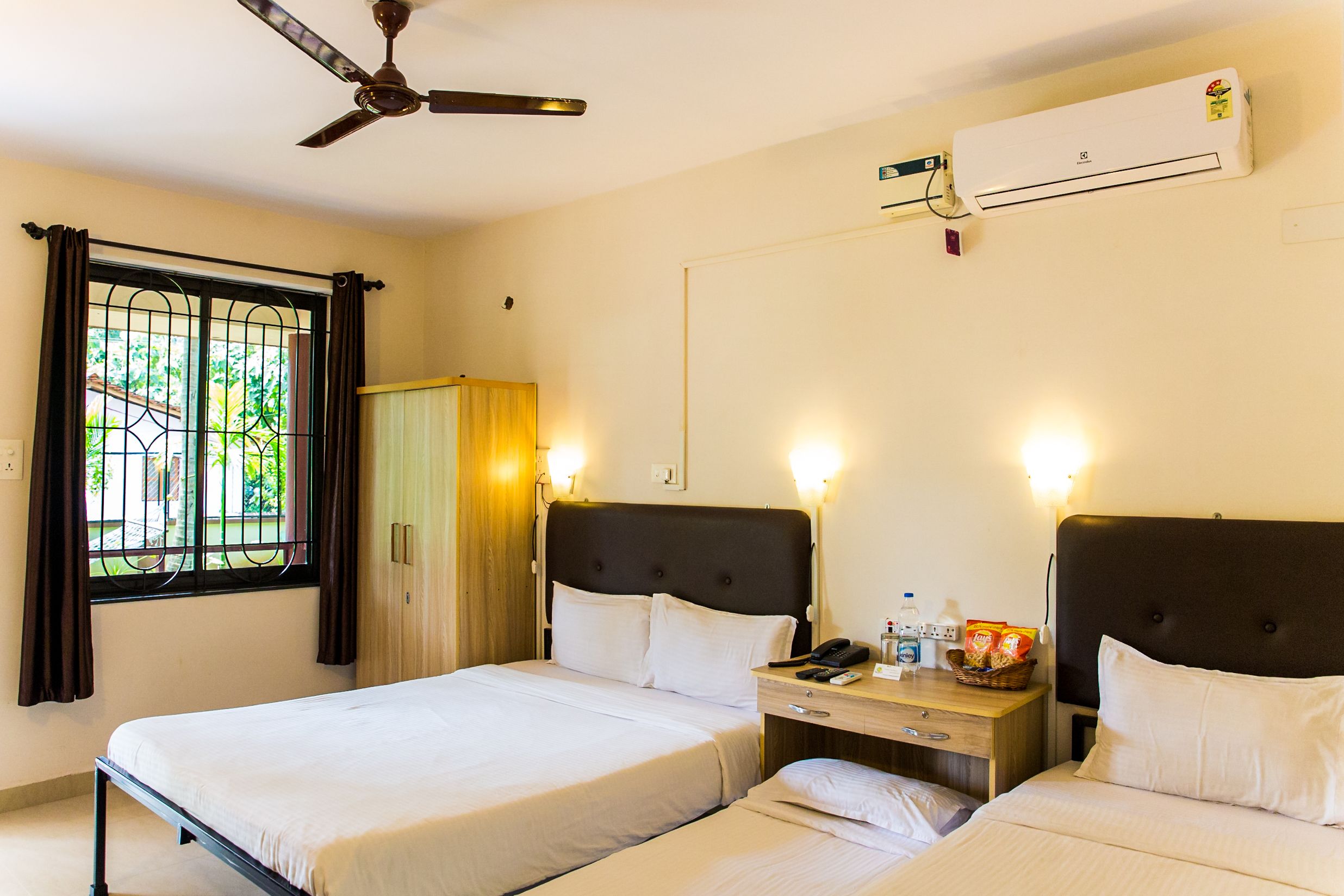 A hotel room with two beds, a ceiling fan, an air conditioning unit, a window with curtains, and a bedside table with a lamp and a basket of snacks.