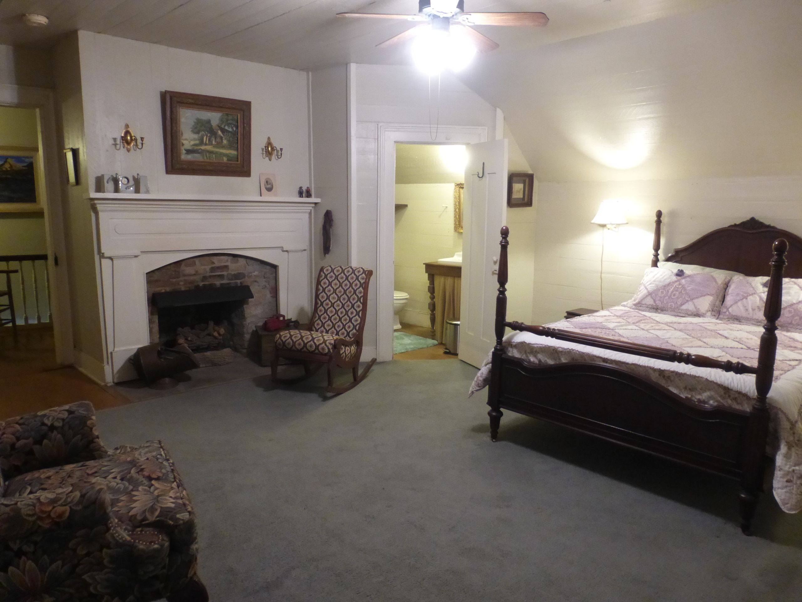 A bedroom with a four-poster bed, a fireplace, a rocking chair, and a carpeted floor.