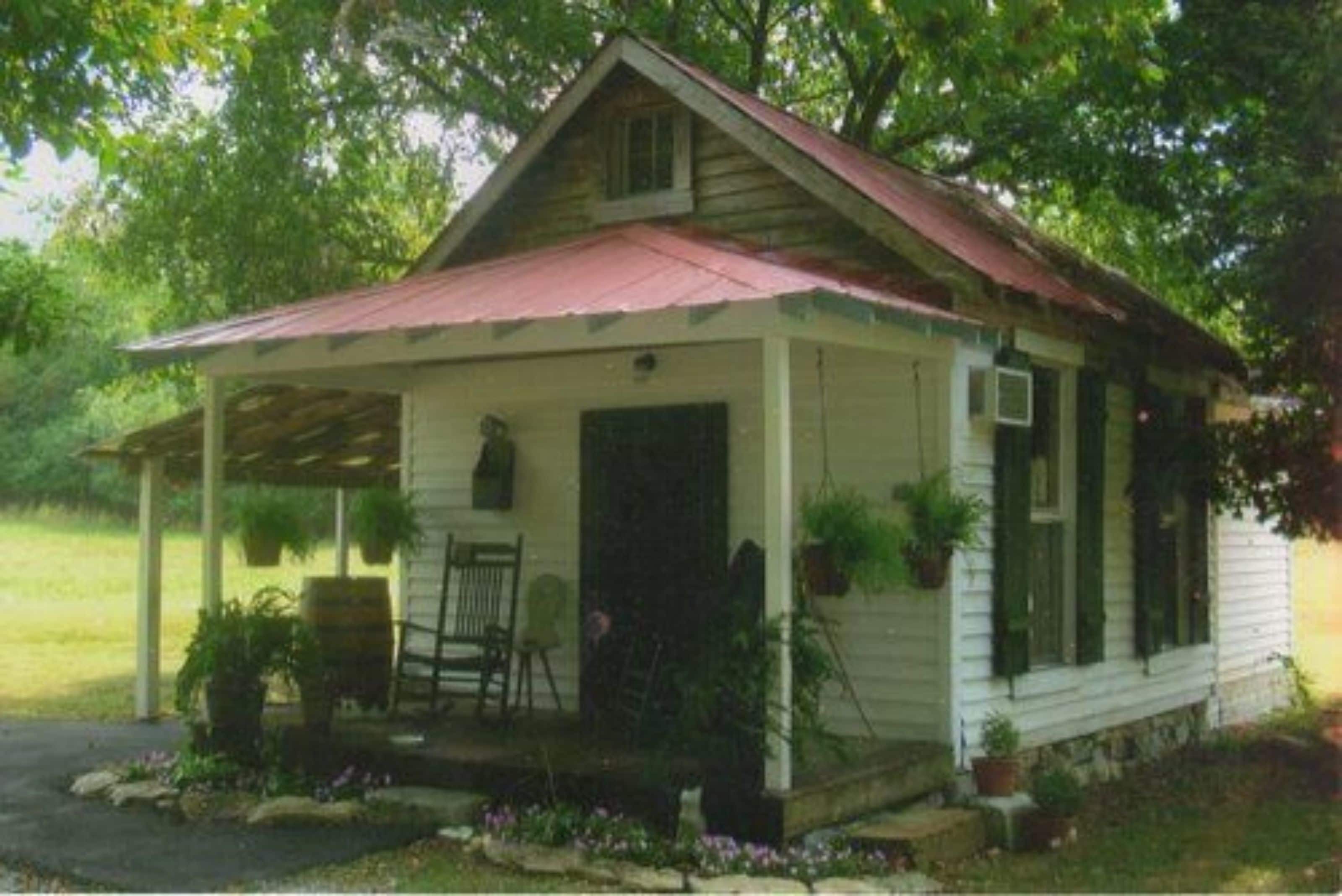 A small, single-story house with a red roof and white siding, featuring a porch with a bench and potted plants.