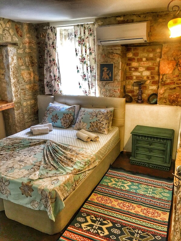 Un dormitorio con cama, cortinas florales, pared de ladrillo, unidad de aire acondicionado y un pequeño aparador verde.