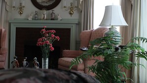 A room with a fireplace, a pink armchair, a table lamp, and a potted fern. - Moore House Bed and Breakfast (Manhattan)