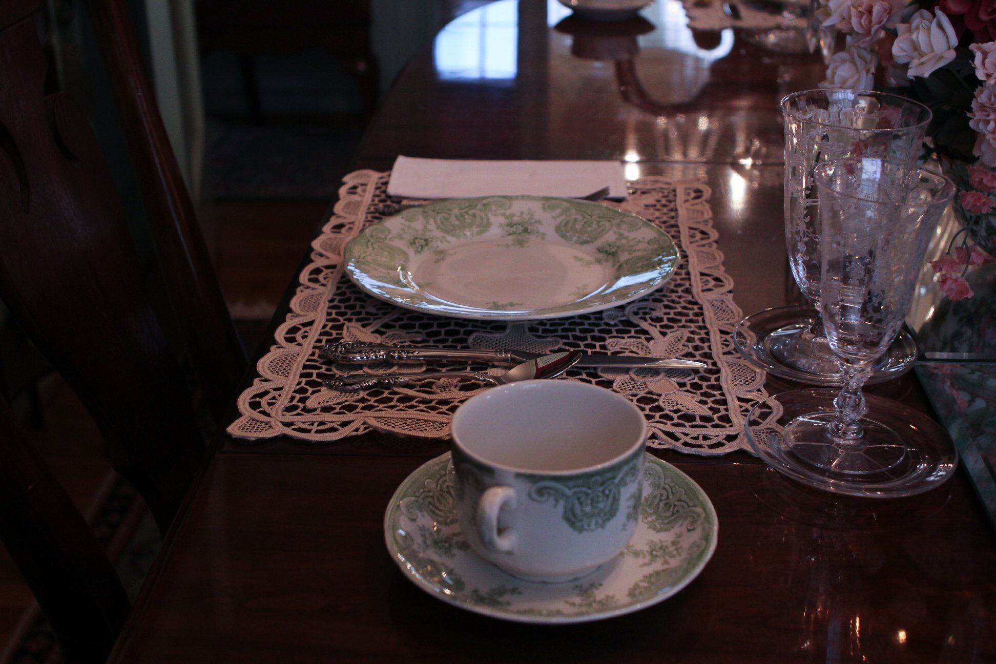 A dining table with a lace tablecloth, a cup and saucer, a glass, and cutlery.